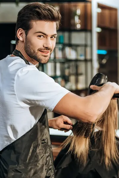 Student in opleiding tot kapper föhnt het haar van een klant in een moderne salon, terwijl hij glimlachend in de camera kijkt. In de spiegel zie je de klant met een tevreden glimlach.